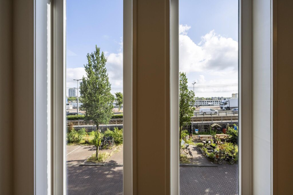 Uitzicht door hoge ramen op De Ruijterkade met bomen, geparkeerde fietsen en een moestuin, met verkeer en spoor op de achtergrond.